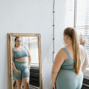 Woman in teal activewear stands before a full-length ornate mirror, checking her reflection in a bright, sunlit room.