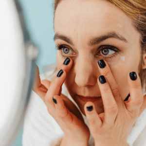 Close-up of a woman applying eye cream under her eyes while looking in a mirror.
